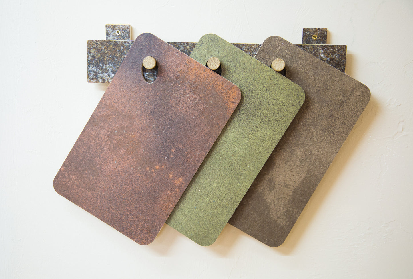 A metallic trio of KAVA trays hanging on three hooks. All are made with coffee waste pressed into birch plywood, the three that hang here together have additional colours added. The first is copper, the second is brass, and the third is bronze.
