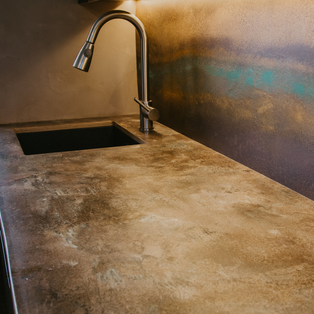 An image of a kitchen worktop with a sink and chrome tap. The worktop is a washed out concrete brown colour, made by the use of spent coffee grounds. The splashback is of a similar finish, containing coffee waste and also oxidised copper powder.